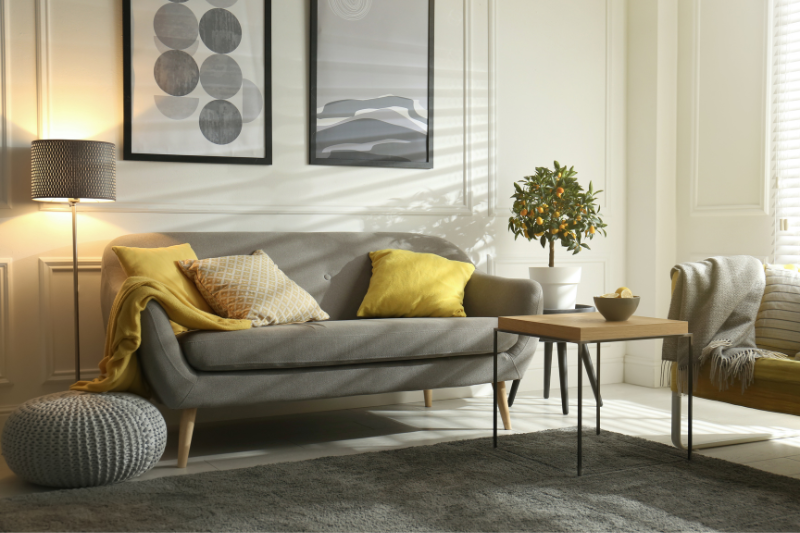 A modern living room with a gray sofa, yellow and gray pillows, a knitted pouf, a wooden coffee table, a potted plant, floor lamp, and abstract wall art. Natural light streams through window blinds.
