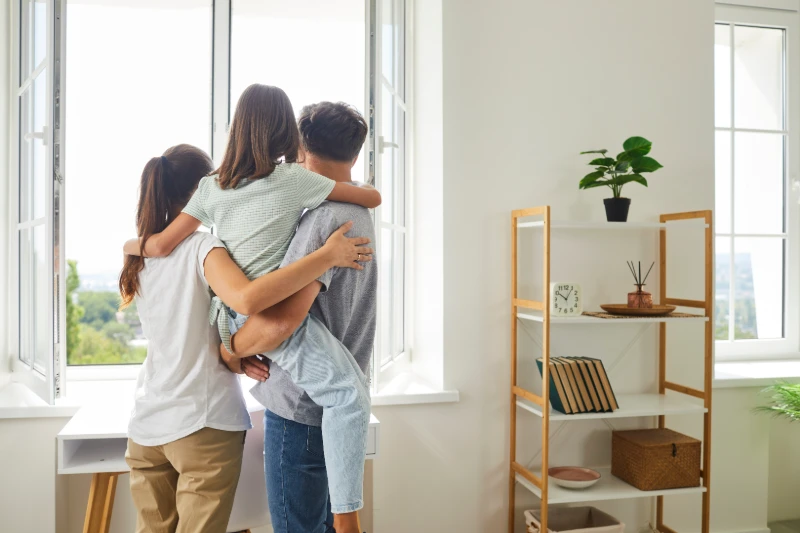 A family of three, seen from behind, stands by an open window. The father holds their child, while the mother stands close. Sunlight fills the bright, tidy room with shelves, a clock, boxes, and a potted plant.