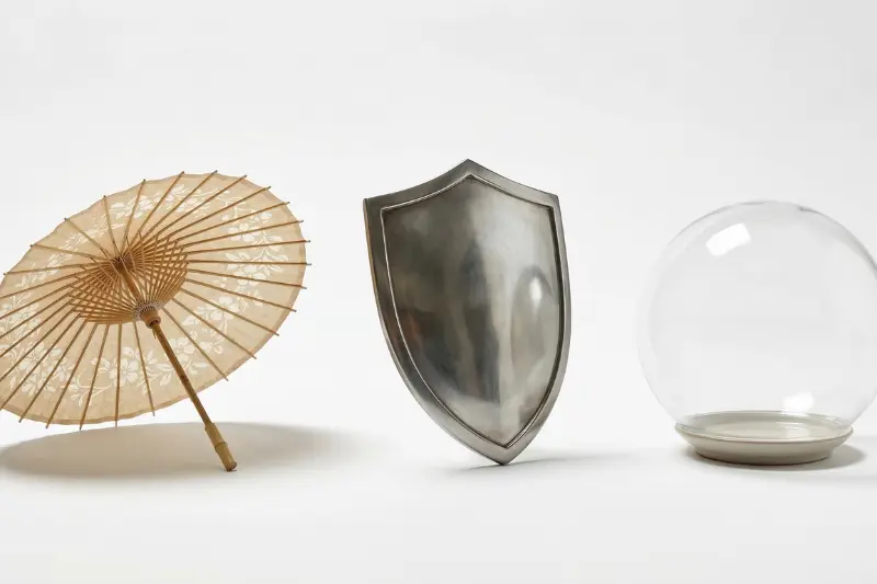 Three protective objects in a row: a paper umbrella, a metal shield, and a glass dome on a white surface.